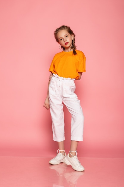 Pełnej długości dziewczynka na różowej ścianie studio. ubrany w pomarańczowy T-shirt, białe spodnie i białe tenisówki.