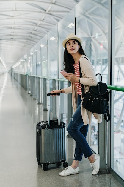 pełnej długości młody student dziewczyna podróżnik trzymając telefon komórkowy relaks na bagażu hali w ręce rozejrzeć się z ciekawą twarz. lady backpacker wyszukująca informacje o locie online na telefonie komórkowym w powietrzu