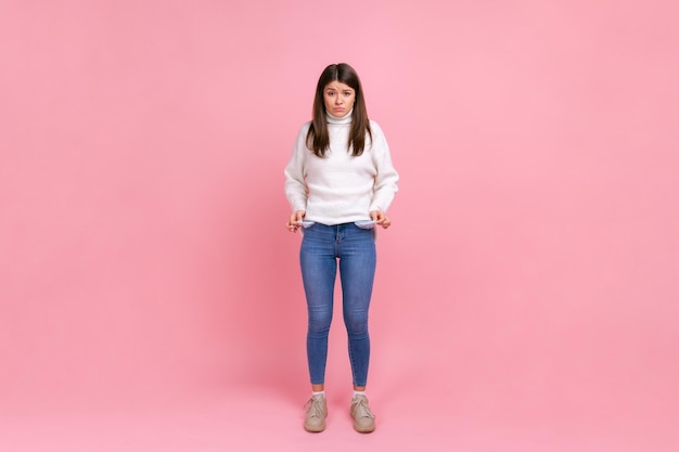 Pełnej długości portret dziewczyny pokazujący puste kieszenie i patrzący sfrustrowany o pożyczki i długi, ubrany w biały sweter w stylu casual. Kryty studio strzał na białym tle na różowym tle.