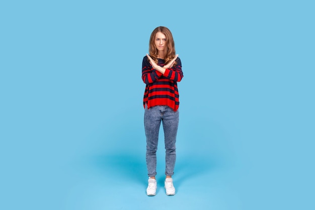 Pełnej długości portret kobiety przekraczania rąk gestem wskazującym ostrzeżenie lub zakaz, co oznacza zatrzymanie zakończenia noszenia sweter w paski w stylu casual Kryty studio strzał na białym tle na niebieskim tle