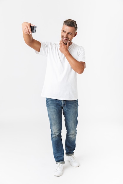 Pełnej Długości Portret Pozytywnego Mężczyzny 30s Ubranych Dorywczo T-shirt I Dżinsy Biorąc Selfie Zdjęcie Na Telefonie Komórkowym, Trzymając W Ręku Na Białym Tle