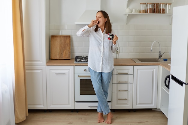 Pełnej długości portret śpiąca kobieta pije gorącą kawę rano, stojąc z filiżanką w rękach i ziewanie, potrzebuje energii, ubrany w białą koszulę i dżinsy.