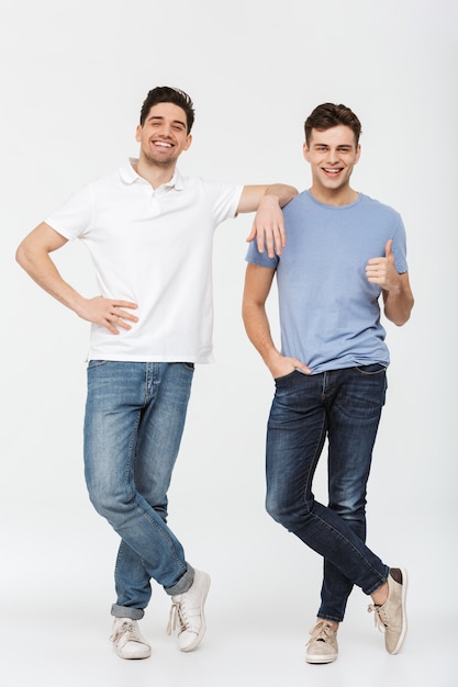 Pełnej długości zdjęcie dwóch przystojnych mężczyzn kumpli 30s na sobie swobodną koszulkę i dżinsy, uśmiechając się i pozowanie razem w aparacie z kciukiem do góry, na białym tle nad białym