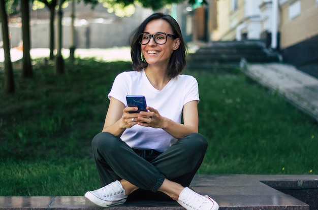 Pełnometrażowe zdjęcie młodej dziewczyny w okularach na twarzy, trzymającej telefon w dłoniach, uśmiechającej się, bawiącej się na dworze.