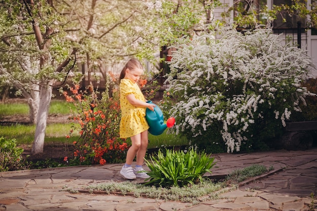 Pełnometrażowy portret dziewczyny w żółtej sukience podlewającej kwiaty z niebieskiego ogrodu konewka...