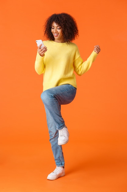 Pełnometrażowy strzał w pionie wesoła urocza technologia z afroamerykańską nowoczesną dziewczyną z afro fryzurą, skakaniem i triumfowaniem jako czytanie świetnych wiadomości ze smartfona, pomarańczowy