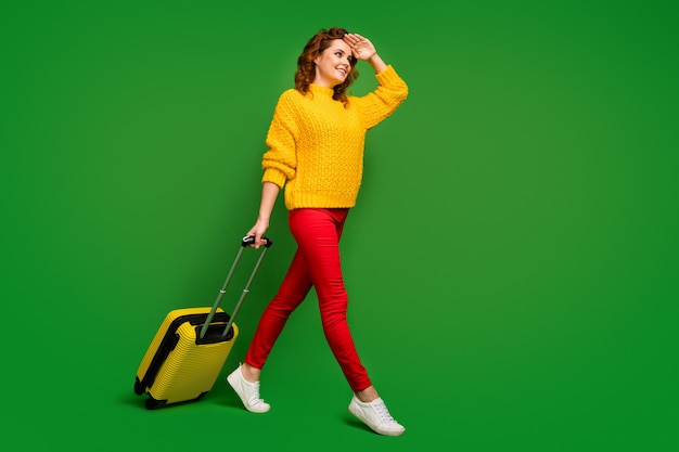 Pełnowymiarowe zdjęcie profilowe ładnej pani tocząca się walizka idź rejestracja spojrzenie daleko dłoń pomóż ukryć oczy przed słońcem nosić żółty sweter czerwone spodnie buty odizolowany zielony kolor ściana