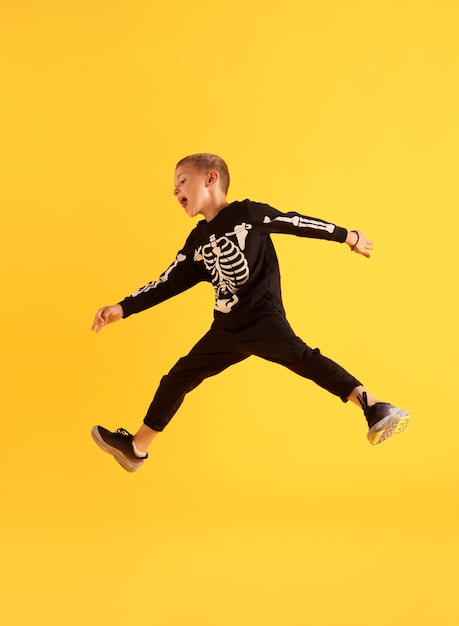 Pełnowymiarowy obraz małego, zabawnego chłopca w czarnym kostiumie skaczącego na żółtym tle studia Gra Koncepcja emocji dzieci z dzieciństwa Impreza Halloween, radość i zabawa, reklama mody