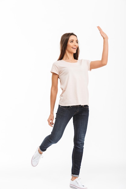 Pełny długość wizerunek Szczęśliwa brunetki kobieta w koszulki odprowadzeniu podczas gdy machający i patrzejący daleko od nad biel ścianą