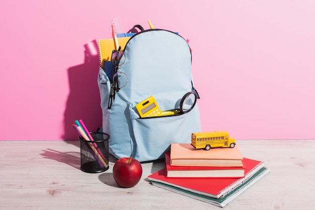 Pełny plecak ze szkolnymi artykułami w pobliżu zabawkowego autobusu szkolnego na książkach dojrzałe jabłko i uchwyt na długopis na różowo