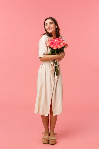 Pełny portret pionowy romantyczna ekstatyczna młoda kobieta otrzymująca wzruszający prezent, obejmująca słodkie bukiety róż, wyglądająca marzycielsko i zachwycona, uśmiechnięta zamyślona, różowa ściana