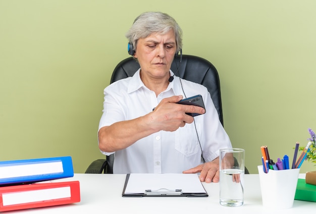 Pewna kaukaska kobieta call center operator na słuchawkach siedzących przy biurku z narzędziami biurowymi trzymającymi i patrzącymi na telefon