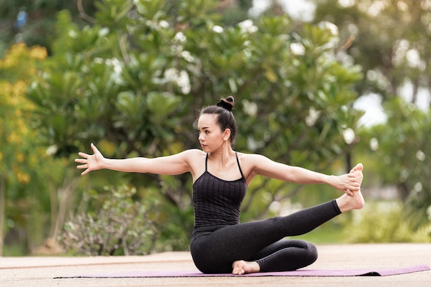 Pewna kobieta w średnim wieku Azjatka w stroju sportowym robi ćwiczenia jogi na macie do jogi na świeżym powietrzu rano na podwórku. Młoda kobieta robi ćwiczenia jogi na świeżym powietrzu w publicznym parku przyrody
