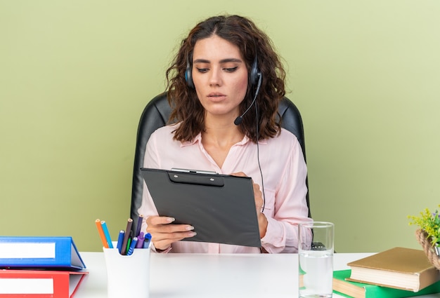 Pewna ładna kaukaska kobieta call center operator na słuchawkach siedzących przy biurku z narzędziami biurowymi piszącymi w schowku na białym tle na zielonej ścianie