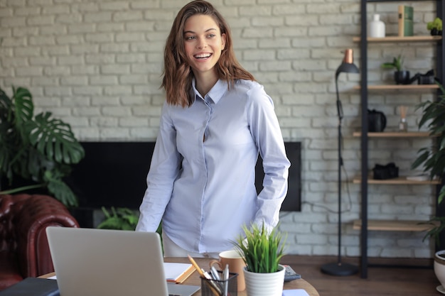 Pewna młoda kobieta z przyjaznym uśmiechem stojąc za biurkiem w domowym biurze patrząc w kamerę.