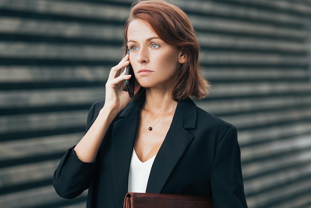 Pewna siebie i poważna bizneswoman z czerwonymi włosami rozmawiająca przez telefon komórkowy
