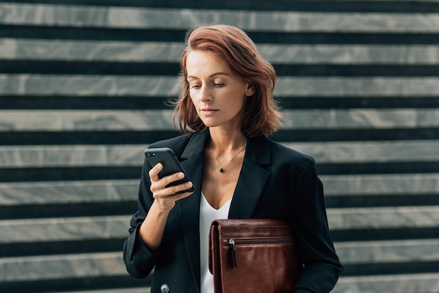 Pewna siebie kobieta w średnim wieku z skórzanym folderem patrząca na swój smartfon, stojąc na zewnątrz