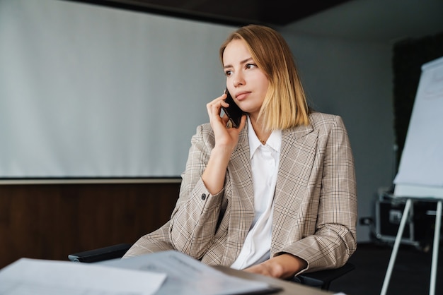 Pewna Siebie Młoda Atrakcyjna Bizneswoman Rozmawia Przez Telefon Komórkowy, Siedząc Przy Biurowym Stole