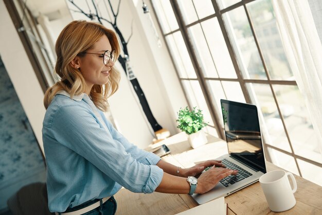 Pewna siebie młoda kobieta korzystająca z laptopa i uśmiechnięta podczas pracy przy oknie