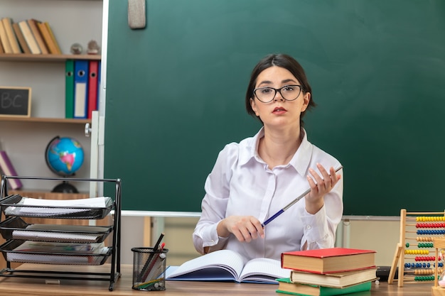 Pewna siebie młoda nauczycielka w okularach trzymająca kij wskaźnikowy siedząca przy stole z narzędziami szkolnymi w klasie