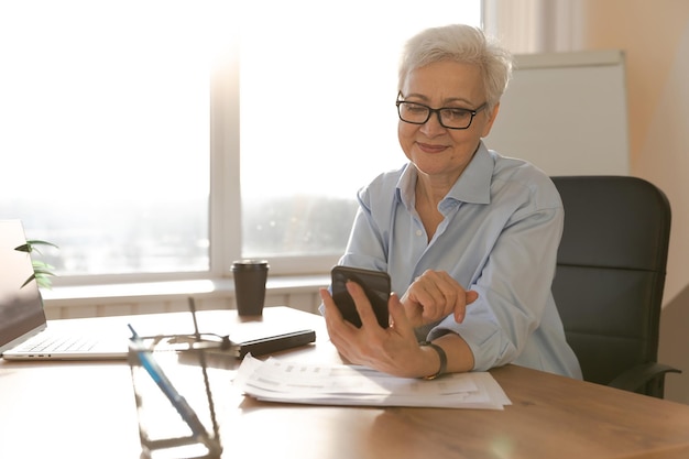 Pewna siebie stylowa europejska starsza kobieta w średnim wieku używająca smartfona w stylowym starszym matu w miejscu pracy