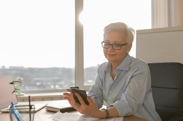Pewna siebie stylowa europejska starsza kobieta w średnim wieku używająca smartfona w stylowym starszym matu w miejscu pracy