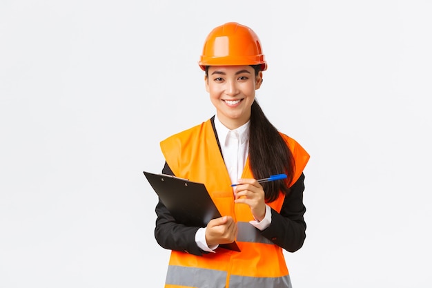 Pewna siebie uśmiechnięta kobieta azjatycka inżynier budowlana, przemysłowa kobieta w kasku ochronnym odwiedza obszar budynku w celu inspekcji, zapisuje notatki w schowku i wygląda na zadowoloną, białe tło
