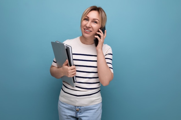 Pewna uśmiechnięta blondynka rozmawiająca na smartfonie trzymająca laptopa w dłoniach na niebiesko