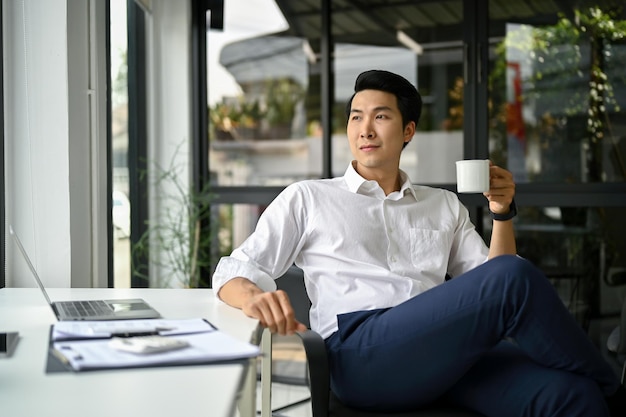 Pewny azjatycki biznesmen siedzi przy biurku w nowoczesnym biurze z filiżanką kawy