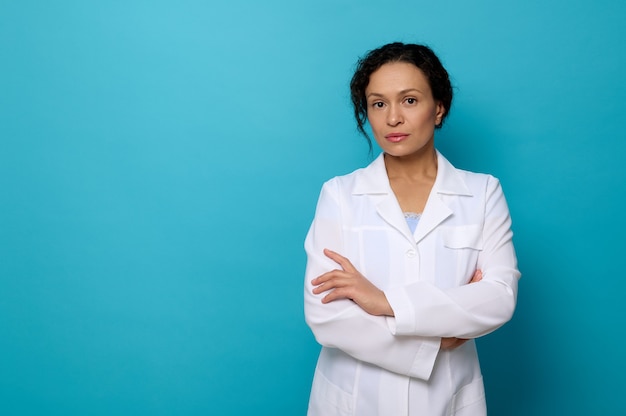 Pewny portret pięknej pogodnej lekarki w białej sukni medycznej, patrząc na kamerę pozującą ze skrzyżowanymi rękami na niebieskim tle z kopią miejsca na reklamę medyczną