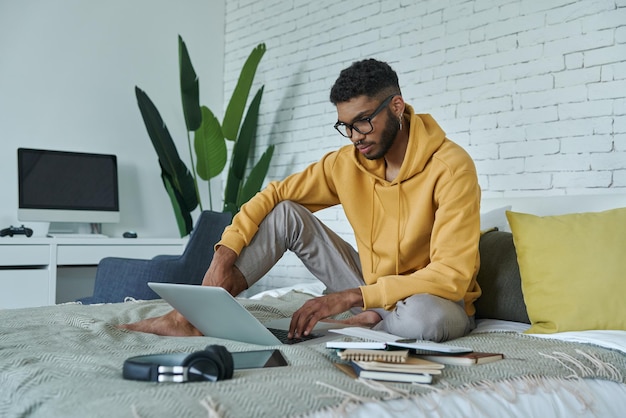 Pewny siebie Afrykanin używający laptopa, siedząc w domu na łóżku