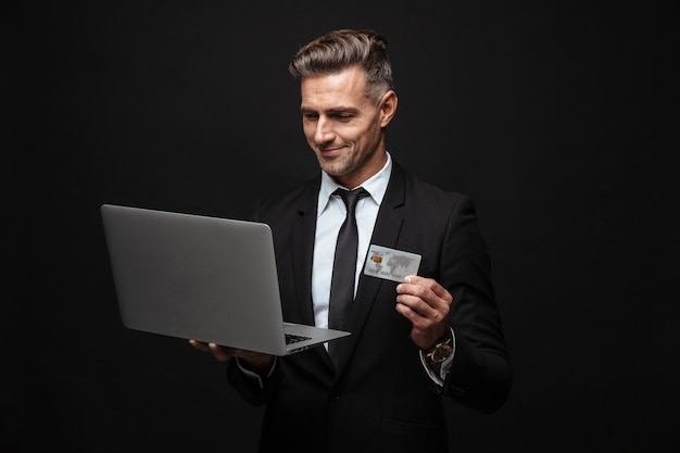 Pewny siebie, atrakcyjny biznesmen w garniturze, stojący na białym tle nad czarną ścianą, używający laptopa, pokazujący plastikową kartę kredytową