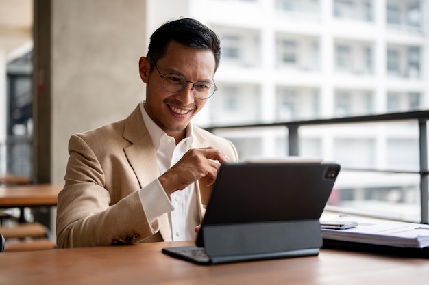 Zdjęcie pewny siebie azjatycki biznesmen siedzi przy stole i pracuje nad swoim cyfrowym tabletem i dokumentem