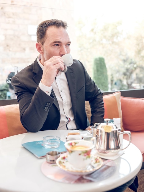 Pewny siebie biznesmen w eleganckim stroju siedzący przy stole na letnim tarasie kawiarni i cieszący się espresso z kubka, patrząc w inną stronę