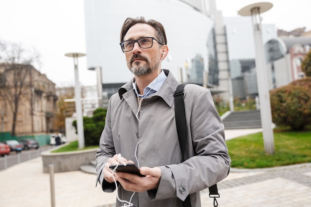 pewny siebie biznesmen w okularach używający telefonu komórkowego i słuchawek podczas spaceru ulicą miasta