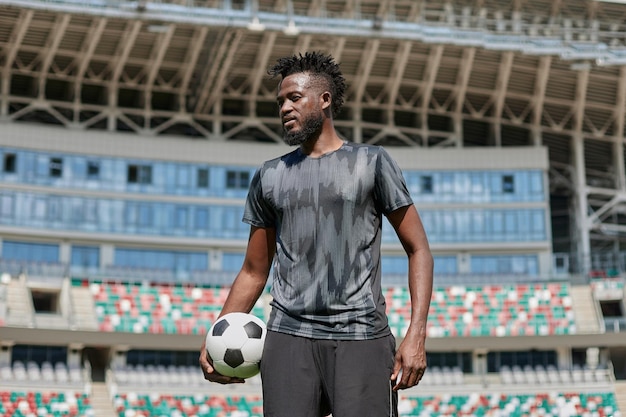 Pewny siebie czarny piłkarz trzymający piłkę w dłoni na stadionie