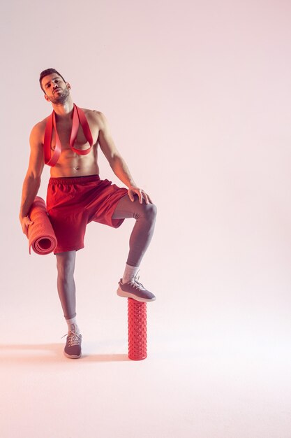 Pewny siebie europejski sportowiec z taśmą oporową, matą fitness i wałkiem do masażu. Młody przystojny brodaty mężczyzna z nagim torsem sportive. Na białym tle na beżowym tle. Sesja studyjna. Skopiuj miejsce