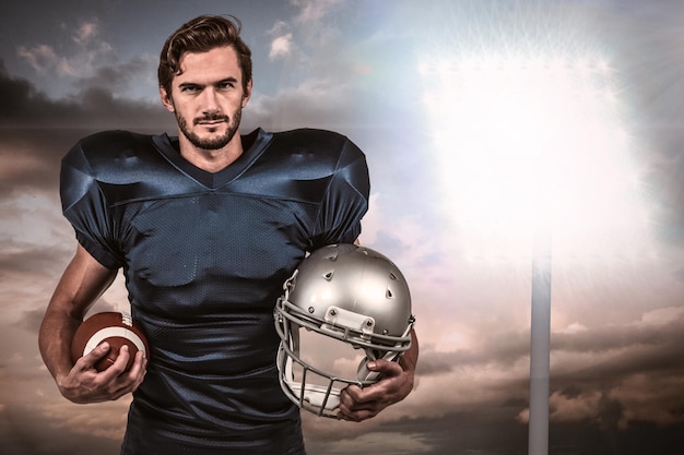 Pewny siebie gracz futbolu amerykańskiego trzymający hełm przed światłem reflektorów na niebie