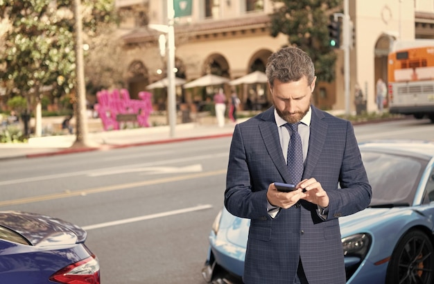 Pewny siebie menedżer wynajmuje online przystojny biznesmen na czacie na smartfonie stoisko przy luksusowym auto odkryty samochód dostępny człowiek w drogim garniturze w pobliżu samochodu ubezpieczenia samochodu biznes udany dealer