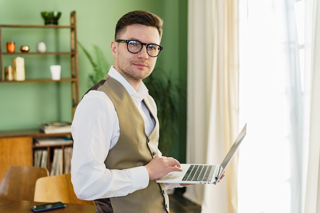 Pewny siebie mężczyzna w okularach i kamizelce stojący i pracujący na laptopie w dobrze oświetlonym pokoju