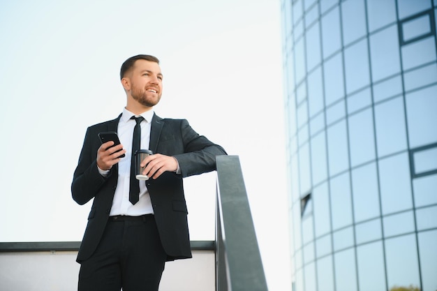 Pewny siebie młody biznesmen używający telefonu komórkowego i pijący kawę w mieście