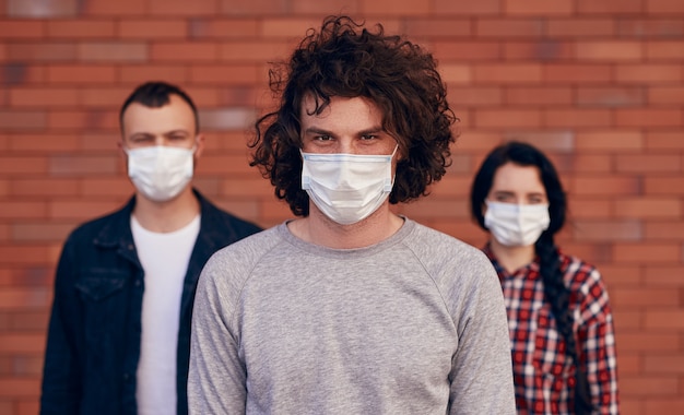 Pewny siebie młody człowiek w masce medycznej, patrząc na kamerę, stojąc w pobliżu przyjaciół podczas epidemii koronawirusa w mieście