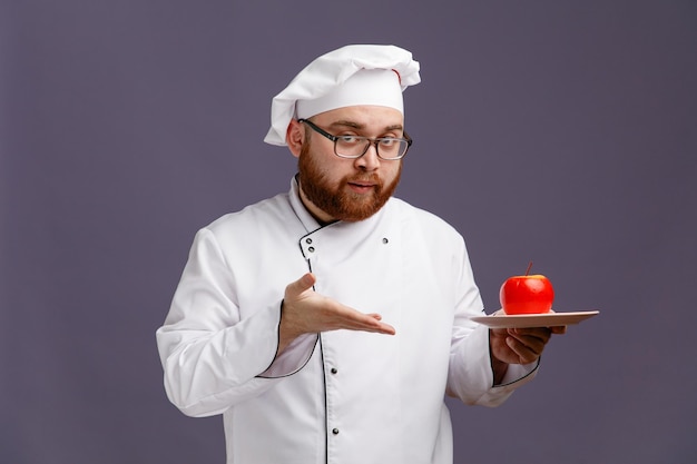 Pewny Siebie Młody Szef Kuchni W Mundurze Okularowym I Czapce, Trzymający Jabłko W Talerzu, Wskazujący Na To Ręką, Patrzącą Na Kamerę Odizolowaną Na Fioletowym Tle