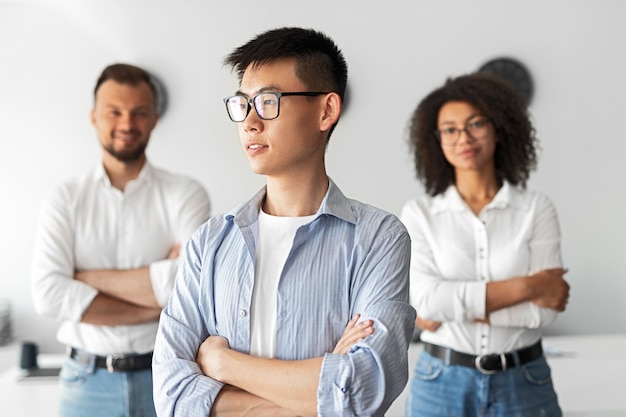 Pewny siebie odnoszący sukcesy młody azjatycki biznesmen w okularach i swobodnym stroju odwracając wzrok stojąc w lekkim biurze z pozytywnymi współpracownikami w tle