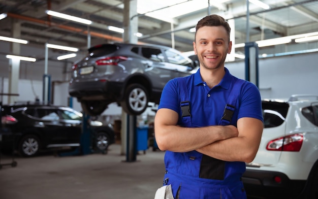 Pewny siebie przystojny młody i doświadczony mechanik samochodowy w kombinezonach roboczych pozujących na tle podnoszonych samochodów w serwisie samochodowym