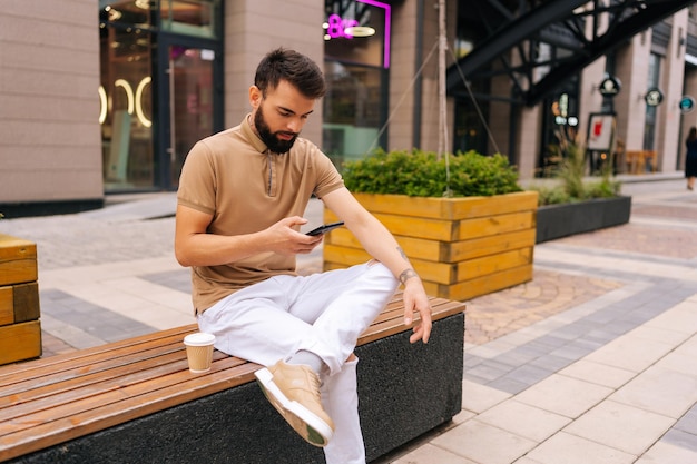 Pewny siebie przystojny młody mężczyzna siedzący na ławce z kawą na wynos i używający smartfona patrzącego na ekran na ulicy miasta