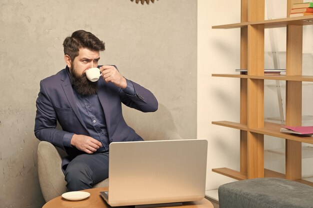 Picie kawy na śniadanie Biznesmen pije kawę w miejscu pracy Brodaty mężczyzna trzyma kubek do picia przy laptopie Ciesz się piciem herbaty lub innego gorącego napoju