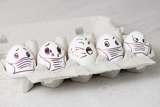 Pięć nieszczęśliwych świątecznych jajek wielkanocnych w maskach. Taca białych jajek z narysowanymi śmiesznymi, zdesperowanymi, płaczliwymi twarzami w maskach medycznych podczas świąt wielkanocnych podczas epidemii koronawirusa z bliska