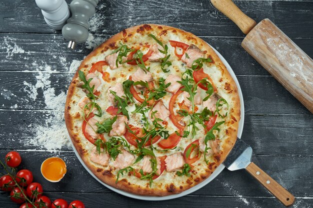 Pieczona pizza z łososiem, pomidorami, serem, białym sosem i rukolą na czarnej drewnianej powierzchni w kompozycji ze składnikami. Widok z góry. Kuchnia włoska