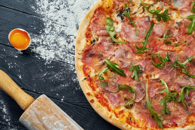 Pieczona pizza z prosciutto z czerwonym sosem i roztopionym serem na czarnym drewnianym stole w kompozycji ze składnikami. Widok z góry